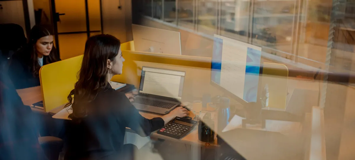 Twee developers aan hun bureau achter een glazen kantoorwand, een van hen werkt op een laptop naast een monitor
