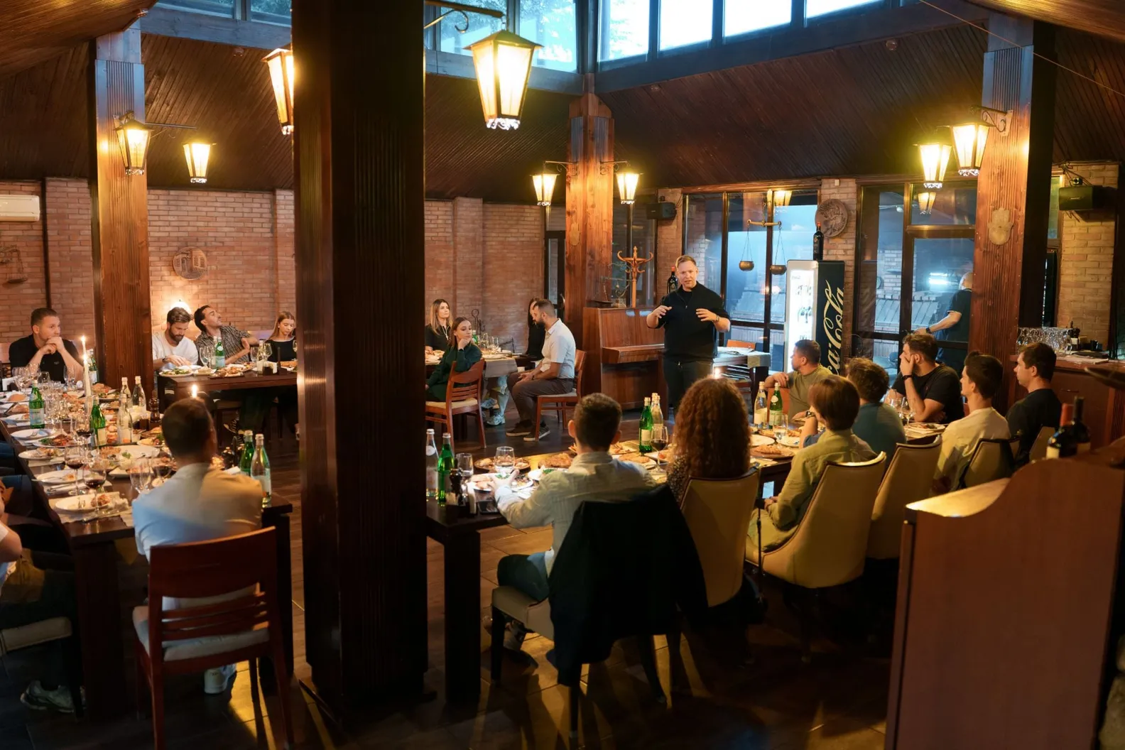 Een eerdere avond van Backstage IT in Moldavië: mensen aan lange tafels in een restaurant met houten balken, bakstenen muren en hangende lantaarns, terwijl een man tussen de tafels staat en de zaal toespreekt.