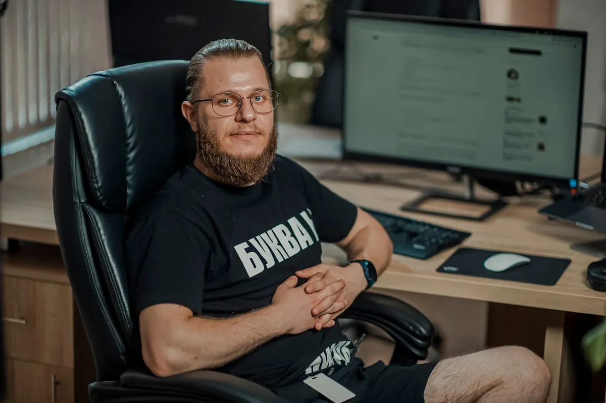Een developer met baard en bril in een zwart T-shirt zit in een leren bureaustoel aan zijn werkplek, met op de achtergrond een monitor met een scherm.