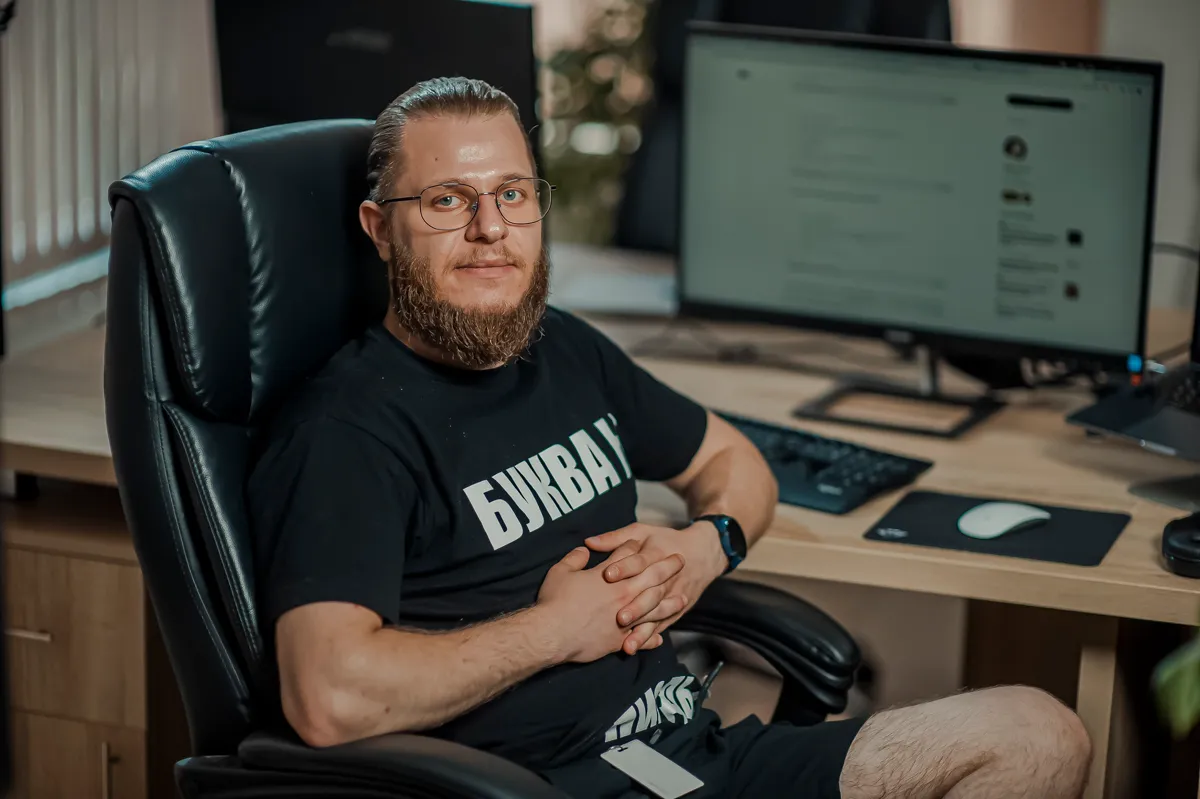 Een softwareontwikkelaar met baard en bril in een zwart T-shirt zit in een leren bureaustoel aan zijn werkplek, met op de achtergrond een monitor met een scherm.