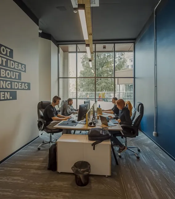 Vier mensen werken aan een bureau bij een groot kantoorraam, naast een muur met de slogan 'It's not about ideas. It's about making ideas happen.'