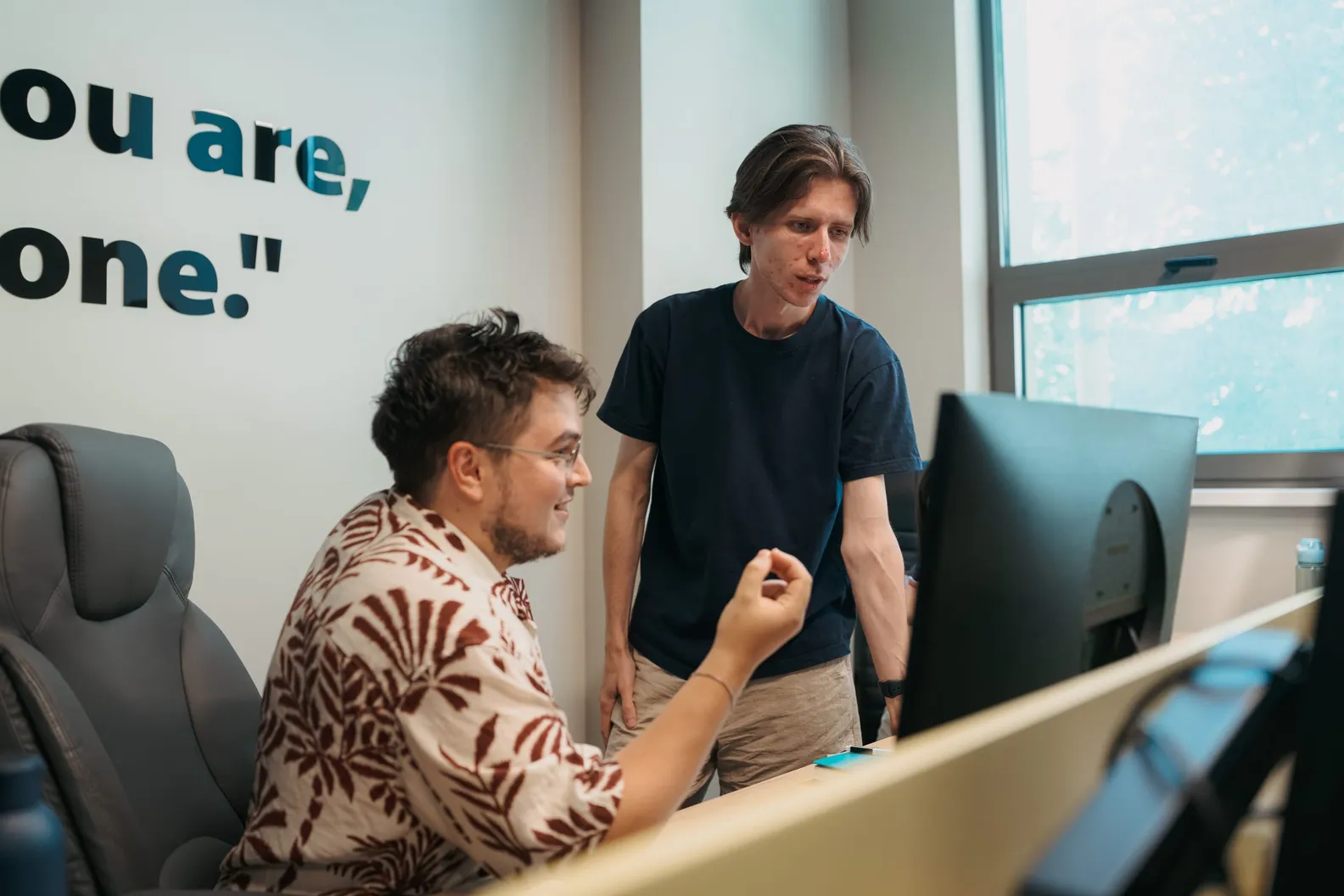 Two developers discussing at a monitor in a bright office, one seated and one standing.