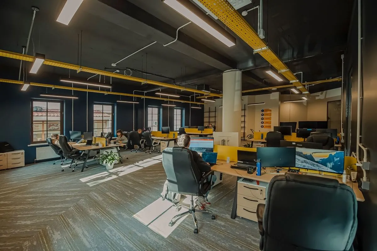 Een open kantoorruimte met gele kabelgoten aan het plafond waar meerdere ontwikkelaars aan bureaus met monitoren werken.