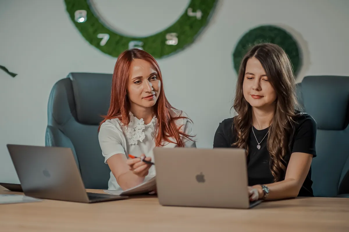 Twee vrouwen werken samen aan een tafel met laptops in een kantoor, waarbij de een tijdens het overleg naar een scherm wijst.
