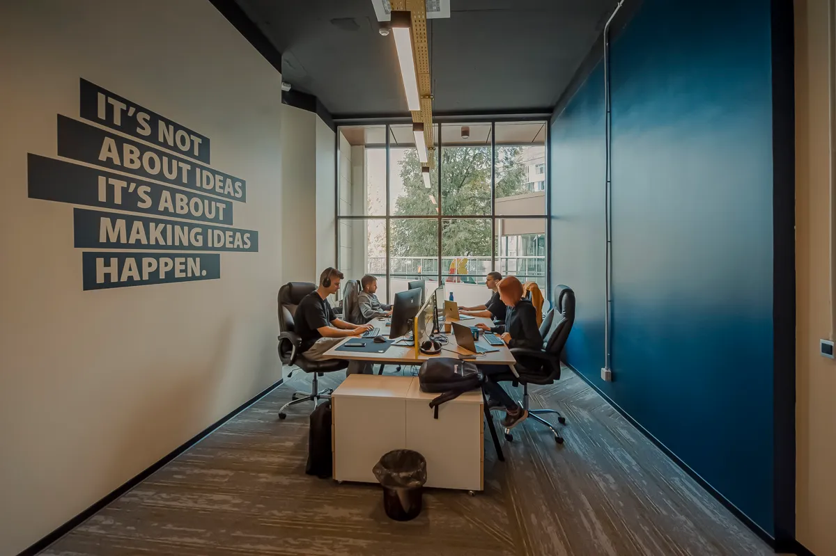 Ontwikkelaars werken aan bureaus bij een raam in een kantoor met op de muur de boodschap dat het draait om ideeën waarmaken.