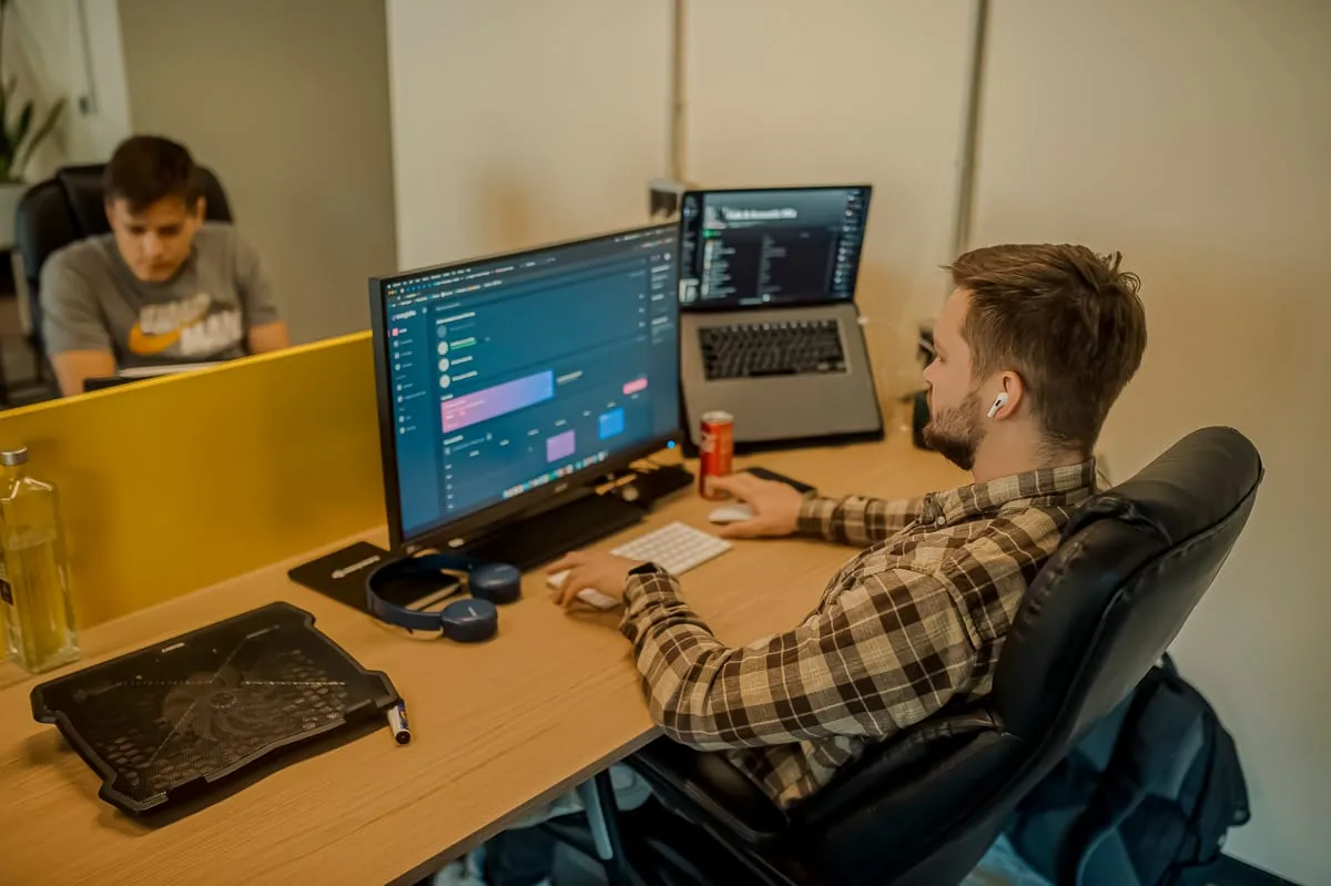Een developer aan zijn bureau werkt aan een webinterface op een groot scherm, met een laptop ernaast en een collega aan het bureau tegenover hem.