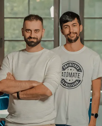 Two team members standing side by side and smiling in front of large windows
