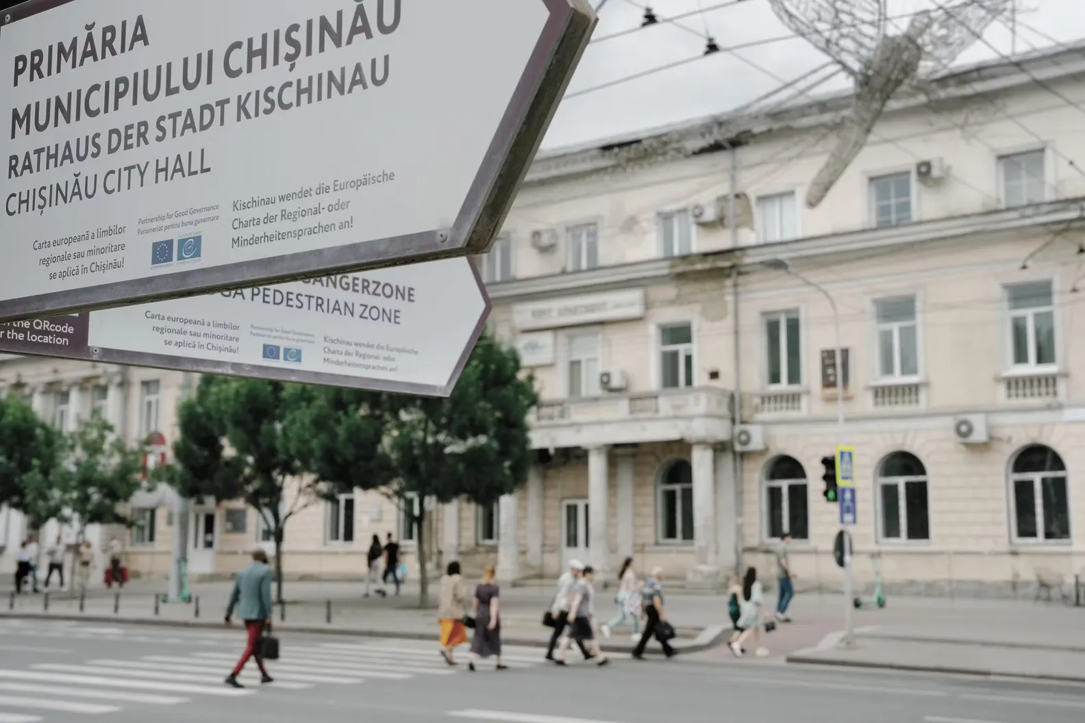 A street sign for Chisinau City Hall in the foreground while pedestrians cross the road in front of a building in central Chisinau.